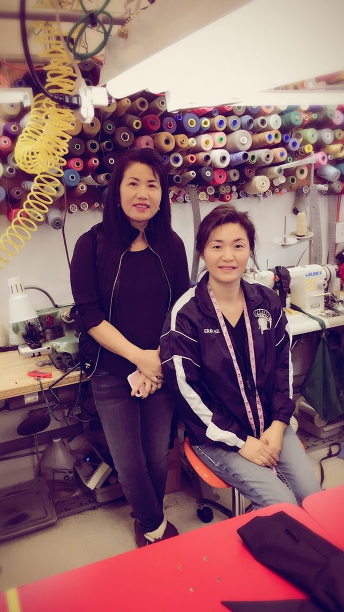 Two women at the alterations bench in front of the thread wall, a tape measure around the neck of the one seated, Juki machines on both sides